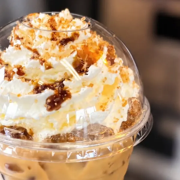 Iced latte topped with whipped cream, caramel drizzle and toffee crumble under a clear dome lid